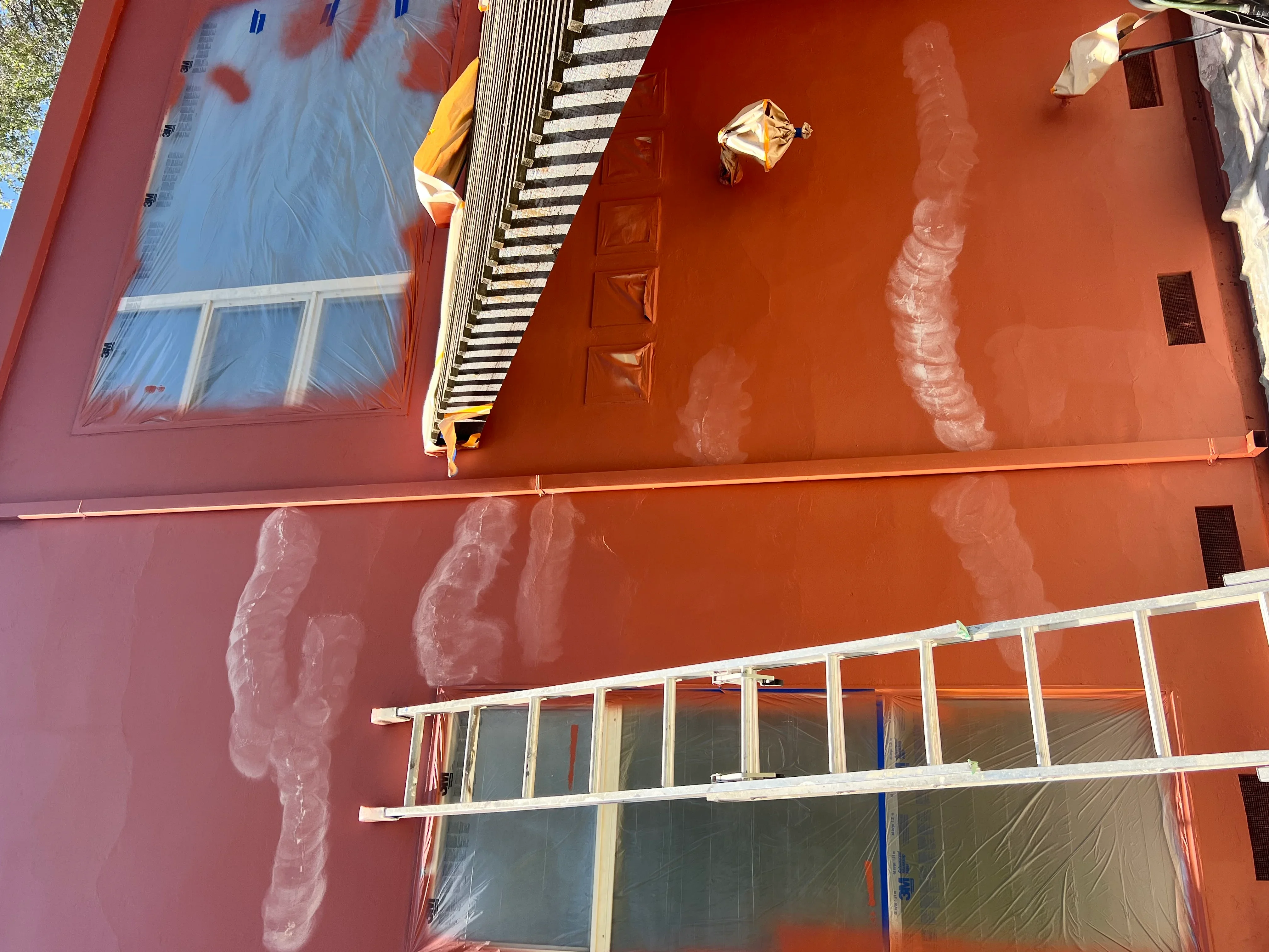 Exterior house painting in progress showing rust-orange painted walls with protective plastic sheeting and scaffolding