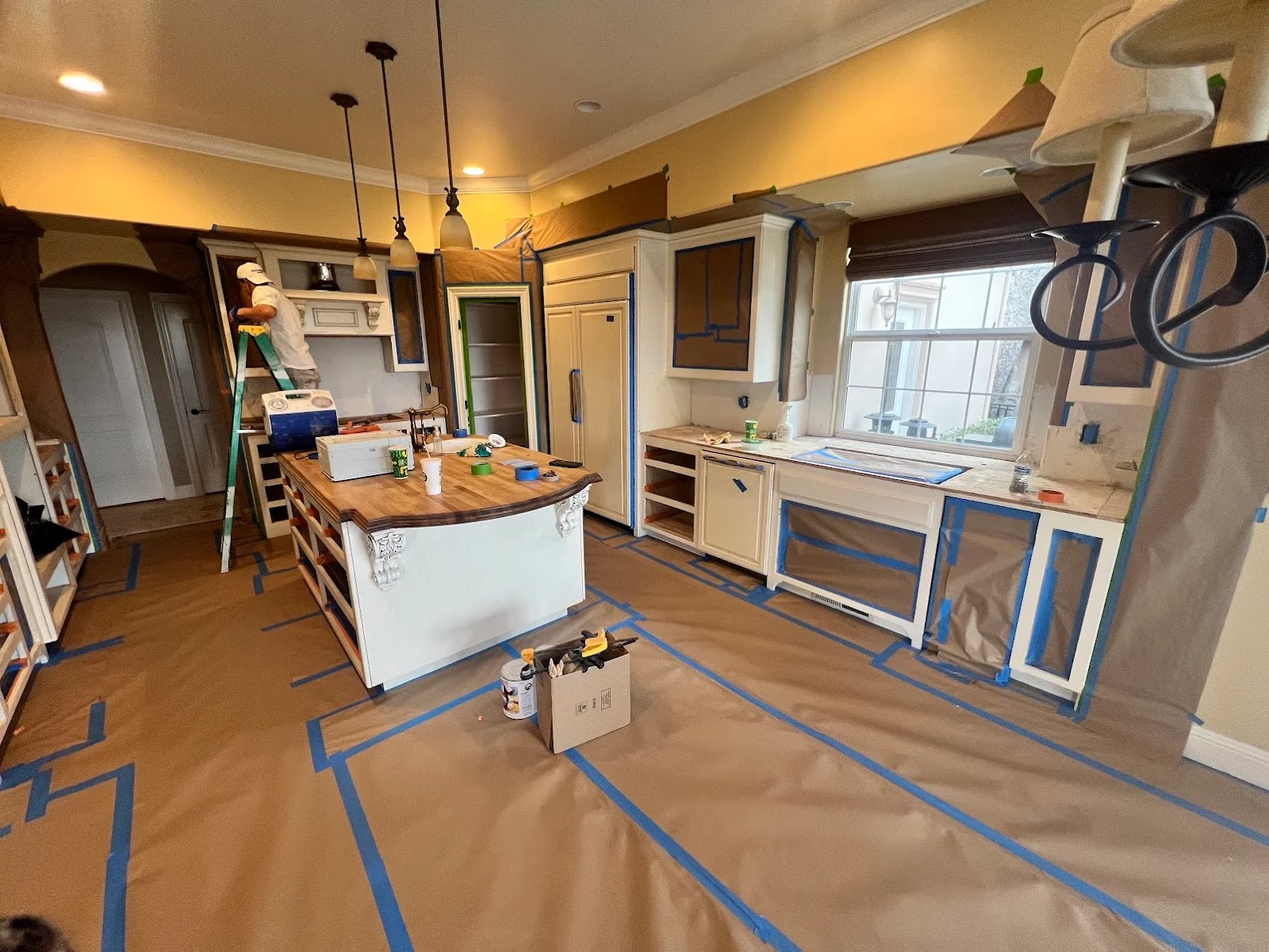 Kitchen interior with protective coverings, blue painter's tape on floors, primed walls, and cream ceiling with recessed lights