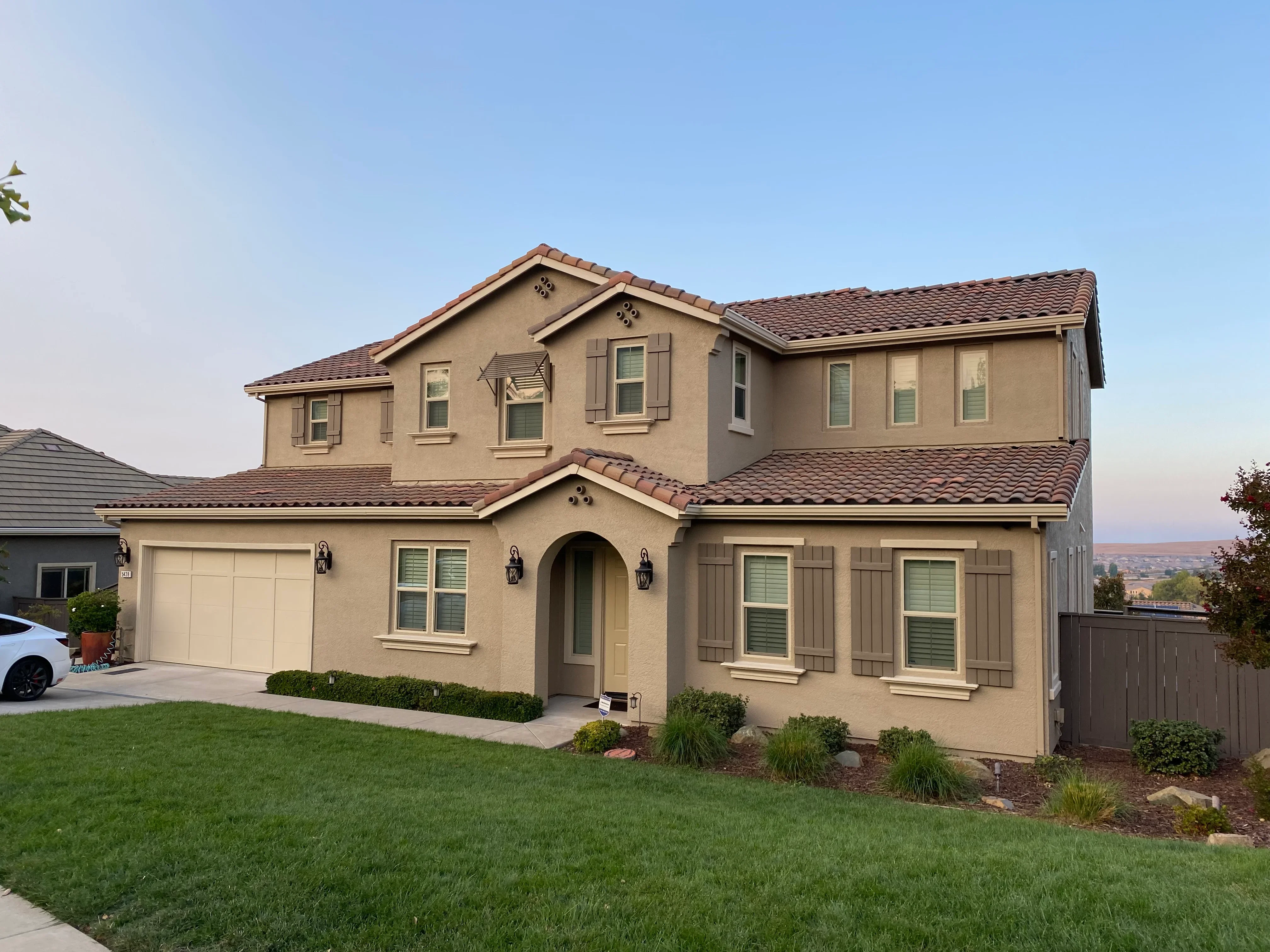 Two-story stucco home with tan exterior paint, brown tile roof, and manicured landscaping
