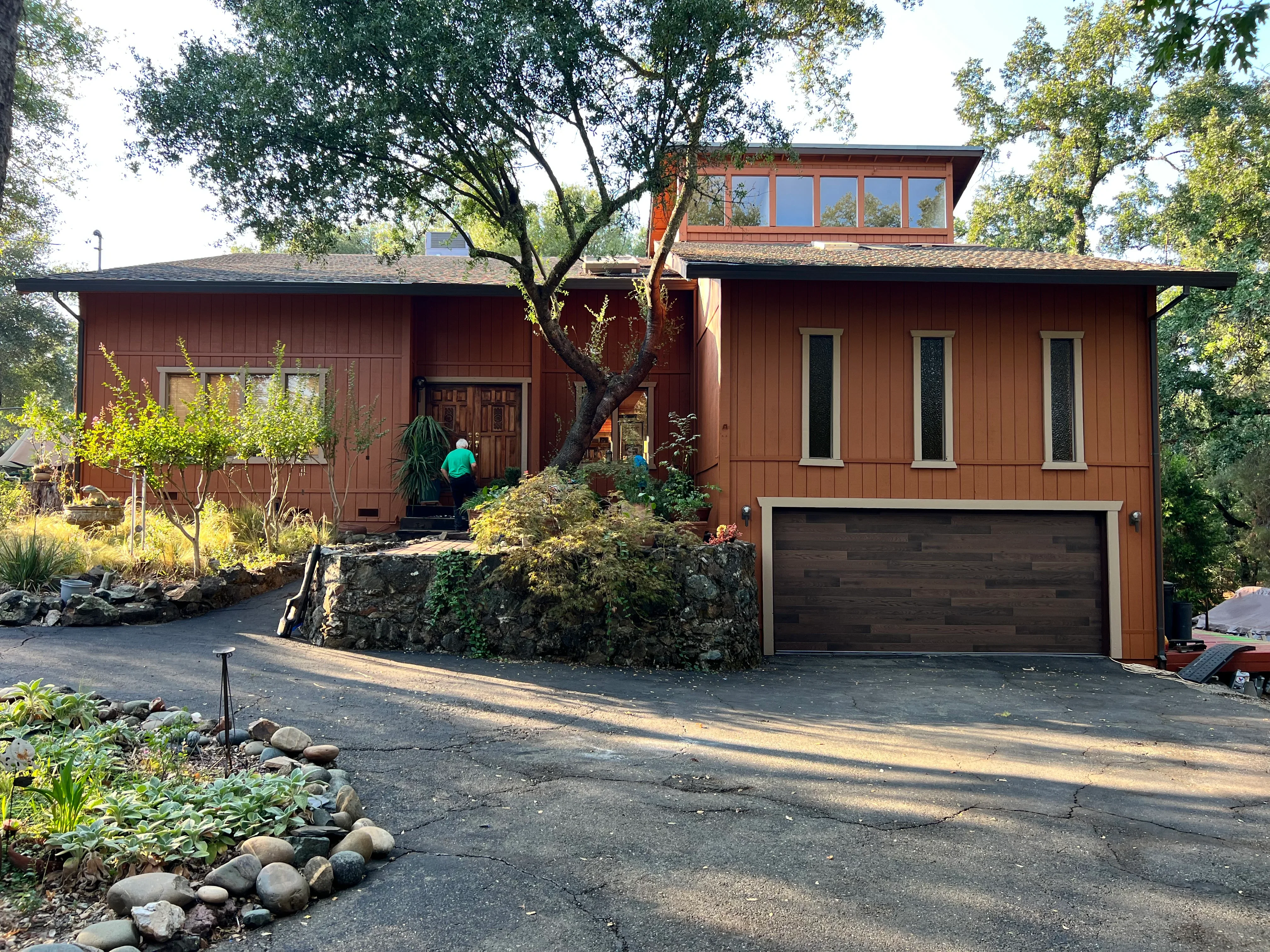 Modern brown home with vertical siding, dark garage door, and mature oak tree in landscaped setting