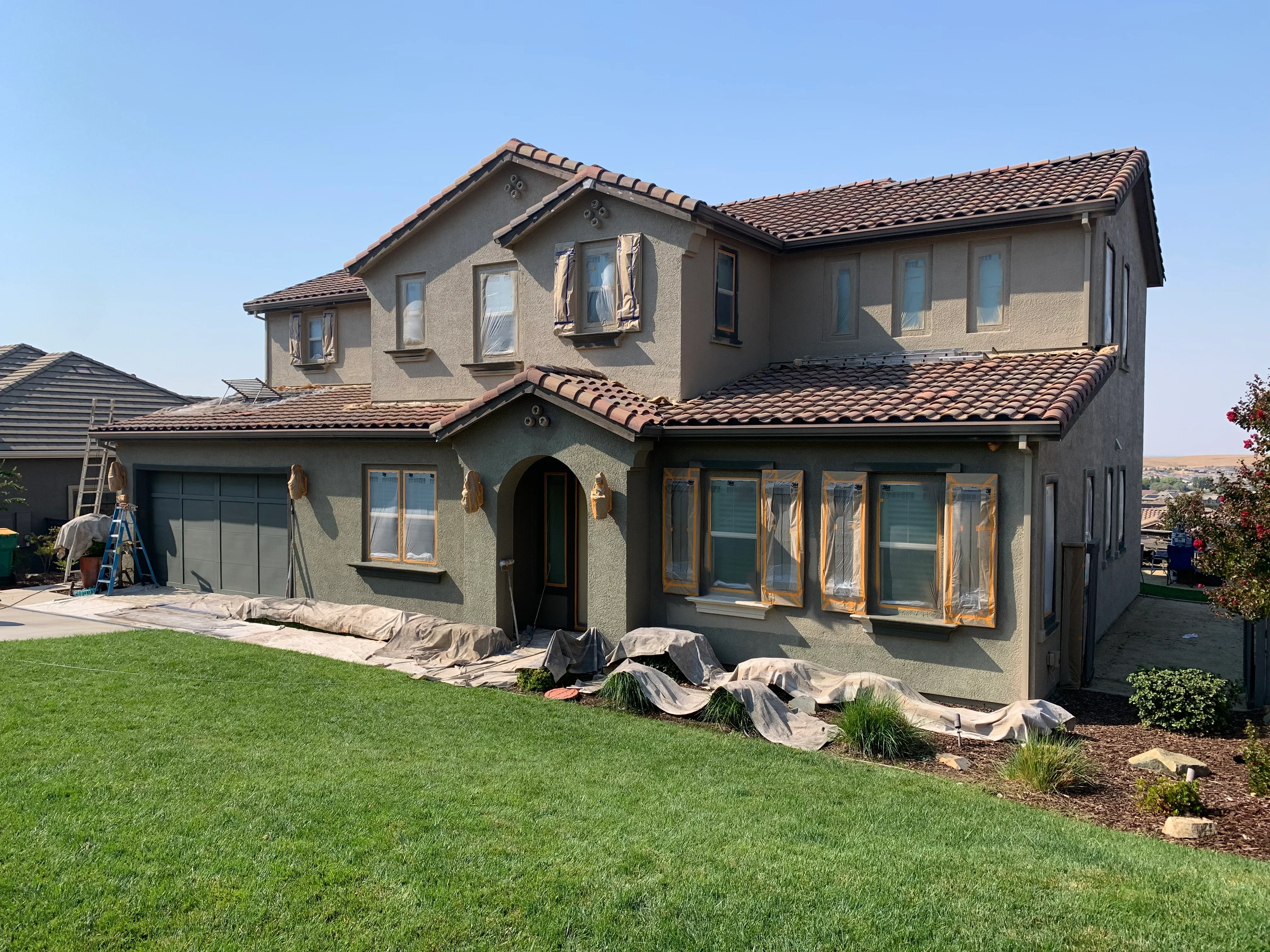 Two-story home with sage green stucco exterior and tan trim during painting project